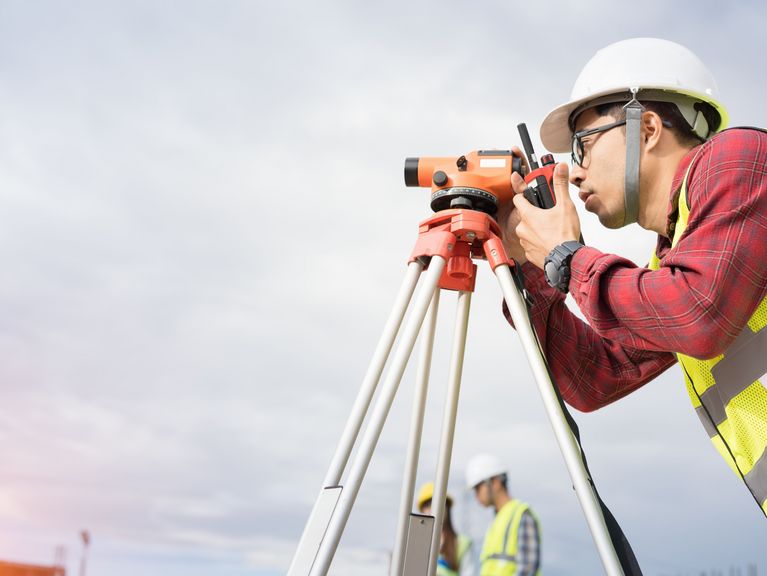 Vermessungsingenieur in Schutzkleidung bedient ein optisches Nivelliergerät auf einem Stativ auf einer Baustelle bei Tageslicht, mit einem weiteren Arbeiter im Hintergrund.