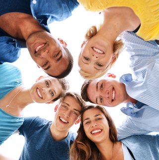 Ein fröhlicher Freundeskreis bildet einen Kreis und blickt von oben in die Kamera, wobei ihre Gesichter ein Bild der Verbundenheit und Freude formen. Die verschiedenen Blautöne ihrer Kleidung kontrastieren schön mit dem hellen Hintergrund, was ein Gefühl von Leichtigkeit und Glück vermittelt.
