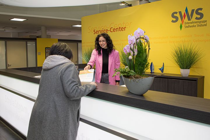 Eine freundliche Angestellte im pinken Mantel bedient eine Kundin am Service-Center der Stadtwerke Stralsund. Im Hintergrund sind Zimmerpflanzen und Auszeichnungen zu sehen sowie die Worte "Vertrauen, Zuverlässigkeit" an der gelben Wand, die die Unternehmenswerte widerspiegeln