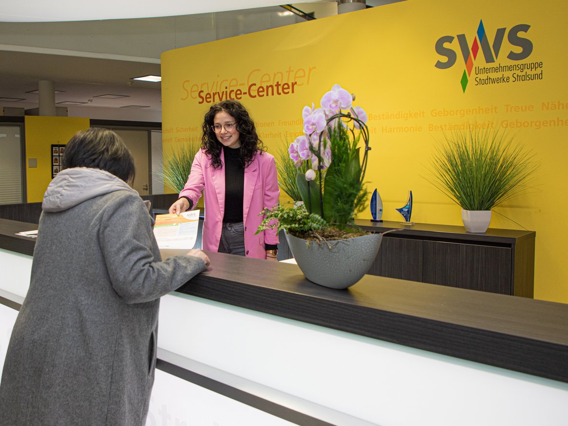 Eine freundliche Angestellte im pinken Mantel bedient eine Kundin am Service-Center der Stadtwerke Stralsund. Im Hintergrund sind Zimmerpflanzen und Auszeichnungen zu sehen sowie die Worte "Vertrauen, Zuverlässigkeit" an der gelben Wand, die die Unternehmenswerte widerspiegeln