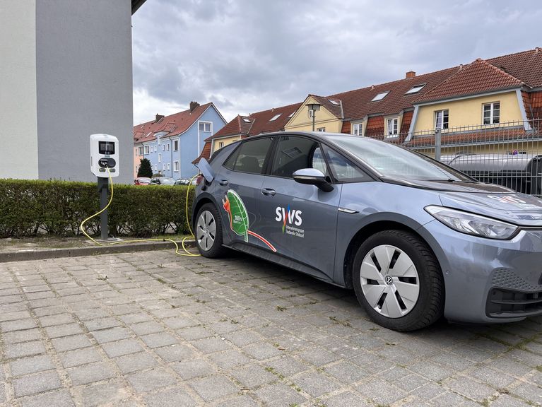 Das Bild zeigt ein graues Elektroauto mit Werbeaufklebern, das auf einem Parkplatz in einem Wohngebiet steht. Im Hintergrund sind Reihenhäuser mit roten Dächern und eine Hecke zu sehen.