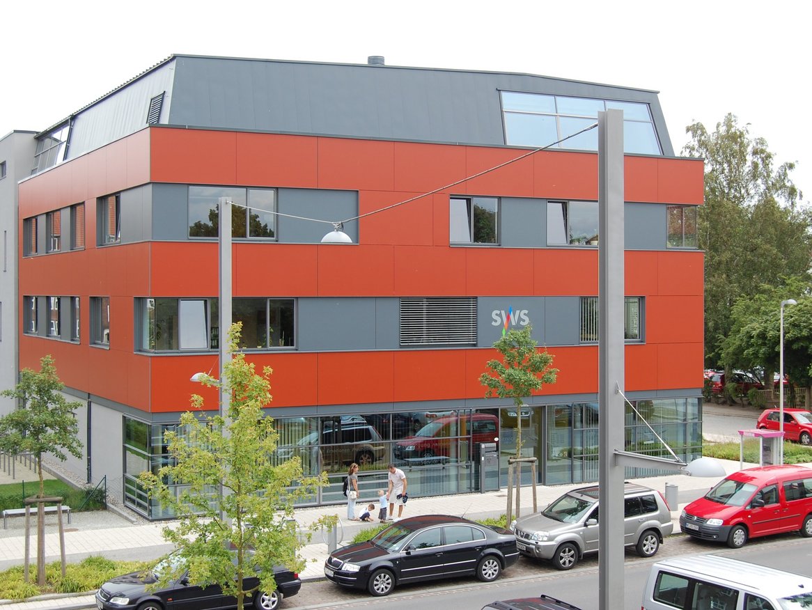 Ein modernes Bürogebäude mit einer auffälligen orangefarbenen Fassade, vor dem parkende Autos zu sehen sind, an einem bewölkten Tag.