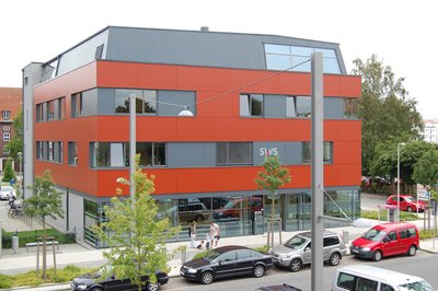 Ein modernes Bürogebäude mit einer auffälligen orangefarbenen Fassade, vor dem parkende Autos zu sehen sind, an einem bewölkten Tag.