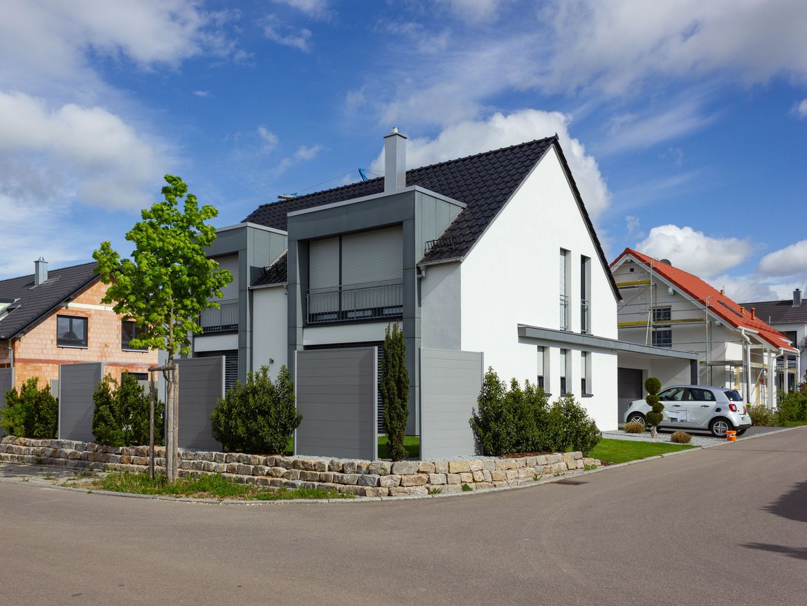 Moderne Wohnhäuser mit sauberen Linien, Giebeldächern und gepflegten Vorgärten in einer ruhigen Vorstadtsiedlung an einem sonnigen Tag mit einigen Wolken am blauen Himmel.