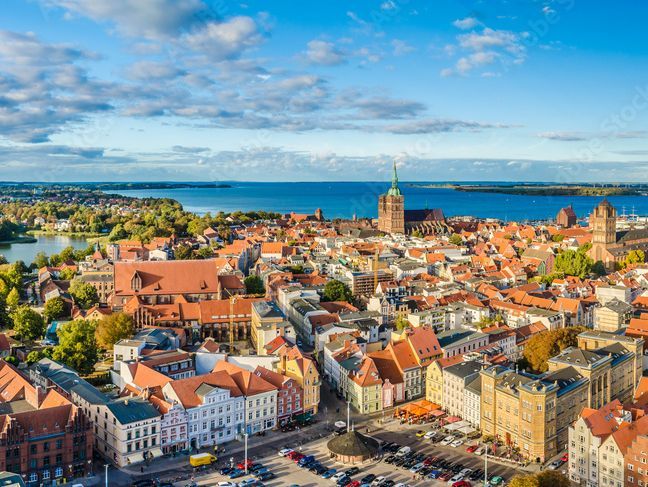 Blick auf die Altstadt von Stralsund mit ihren charakteristischen roten Dächern und historischen Gebäuden. Im Hintergrund ist das blaue Wasser des Strelasunds zu sehen, das die Stadt vom Festland trennt, was die malerische Lage Stralsunds an der Ostseeküste unterstreicht.