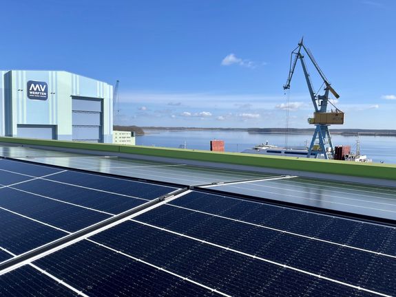 Eine große Solaranlage im Vordergrund mit einem Blick auf eine Wasserlandschaft und Industrieanlagen im Hintergrund, unter einem klaren Himmel – ein Bild, das die Integration von erneuerbaren Energien in industriellen Zonen illustriert.