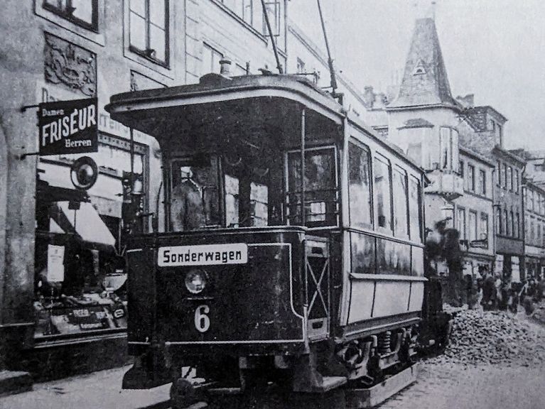 Ein historisches Schwarz-Weiß-Foto einer Straßenbahn der Linie 6, die durch eine belebte Straße mit klassischer Architektur fährt.