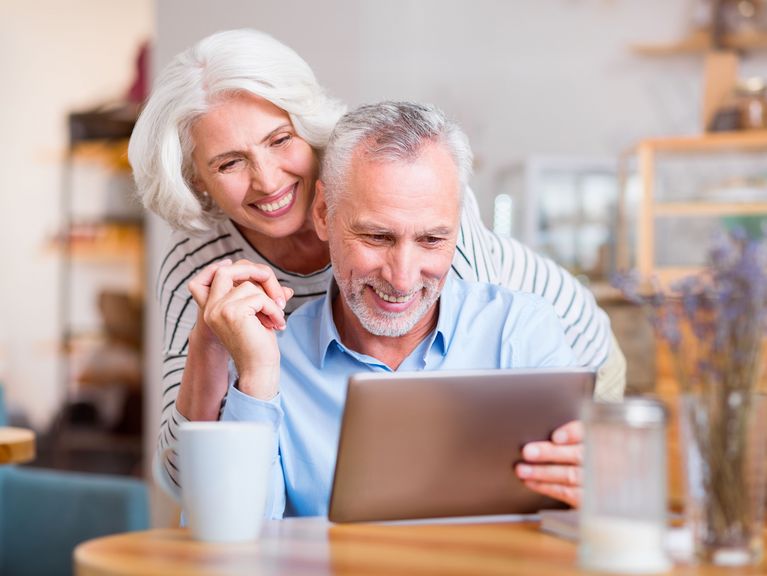 Ein älteres Paar, sichtlich in trauter Zweisamkeit, schaut gemeinsam auf ein Tablet. Sie lächeln fröhlich und verbunden, ein Bild häuslicher Harmonie und moderner Techniknutzung im Alter.