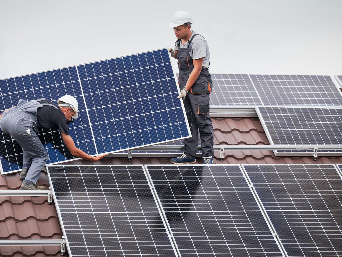 Zwei Monteure in Arbeitskleidung und Schutzhelmen installieren sorgfältig Solarpaneele auf einem Hausdach. Diese Szene hebt das Engagement für erneuerbare Energien und die Wichtigkeit der Solarenergie für eine nachhaltige Zukunft hervor.